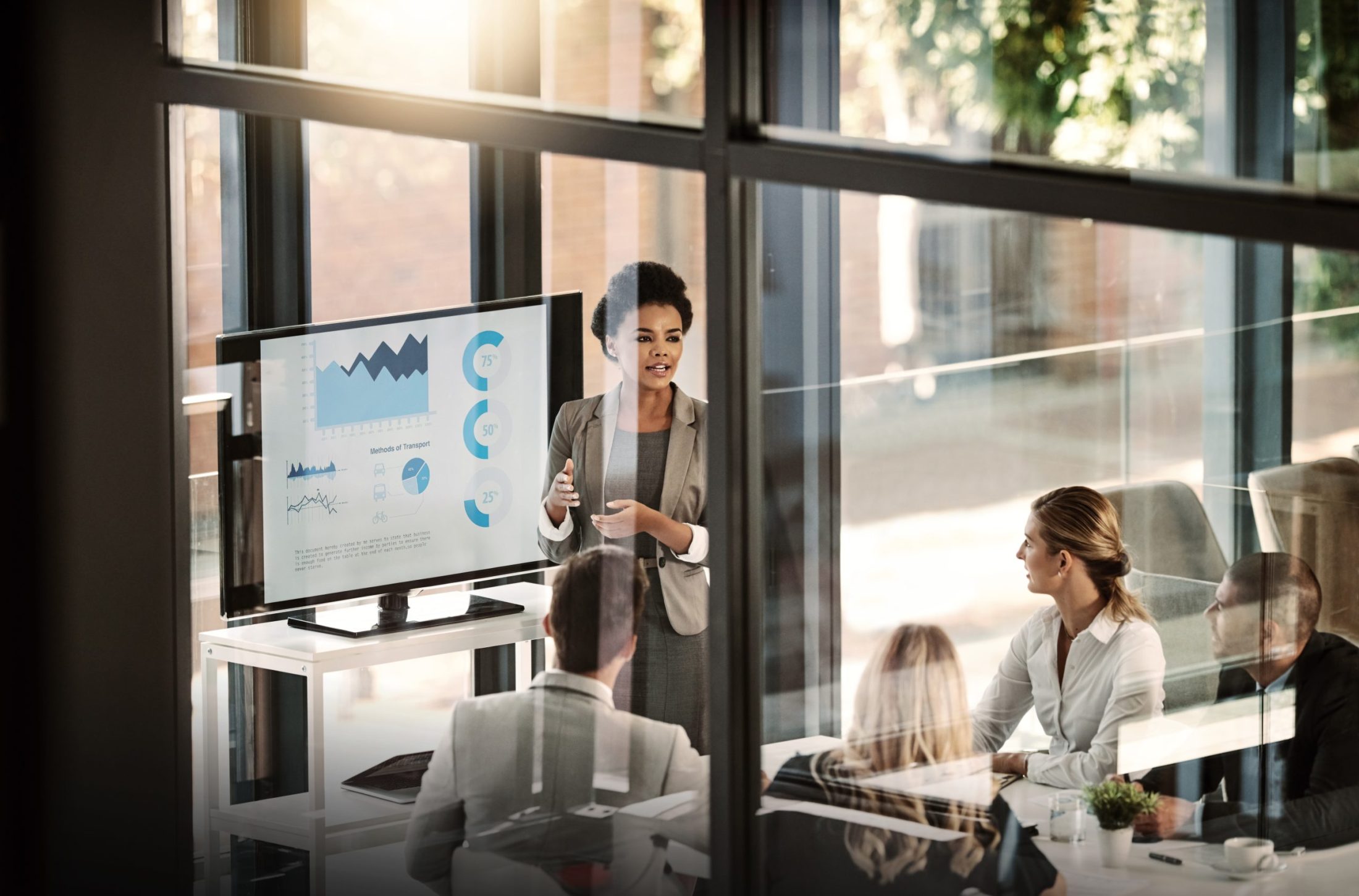 A woman presents charts on a screen to four colleagues seated around a table in a glass-walled office.