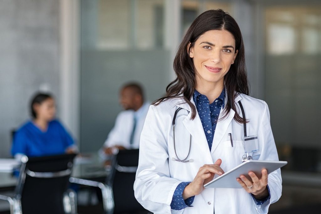 A doctor in a white lab coat checking a tablet and smiling.