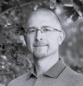 A man with glasses and a short beard, wearing a collared shirt, stands outdoors in front of blurred foliage. The image is in black and white.