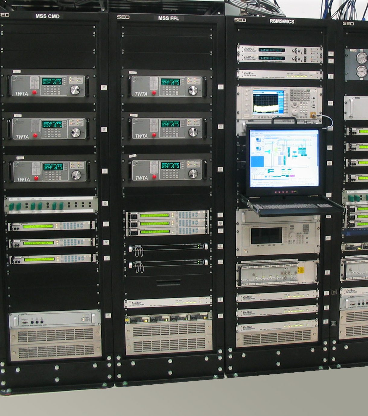 Un rack de serveur avec plusieurs panneaux d'équipements électroniques, notamment des générateurs de signaux, des analyseurs de spectre et des dispositifs de surveillance, avec un ordinateur portable affichant les données devant.