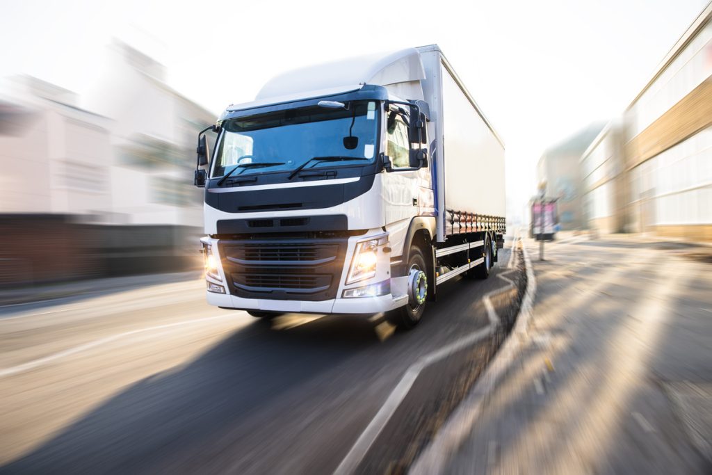 A large white truck with a trailer is driving quickly on a city street with blurred surroundings, indicating motion. The background includes buildings and another vehicle in the distance.