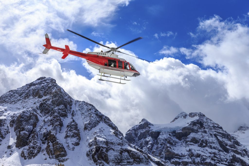 A red rescue helicopter flying in front of snow-covered mountains under a blue sky with clouds.