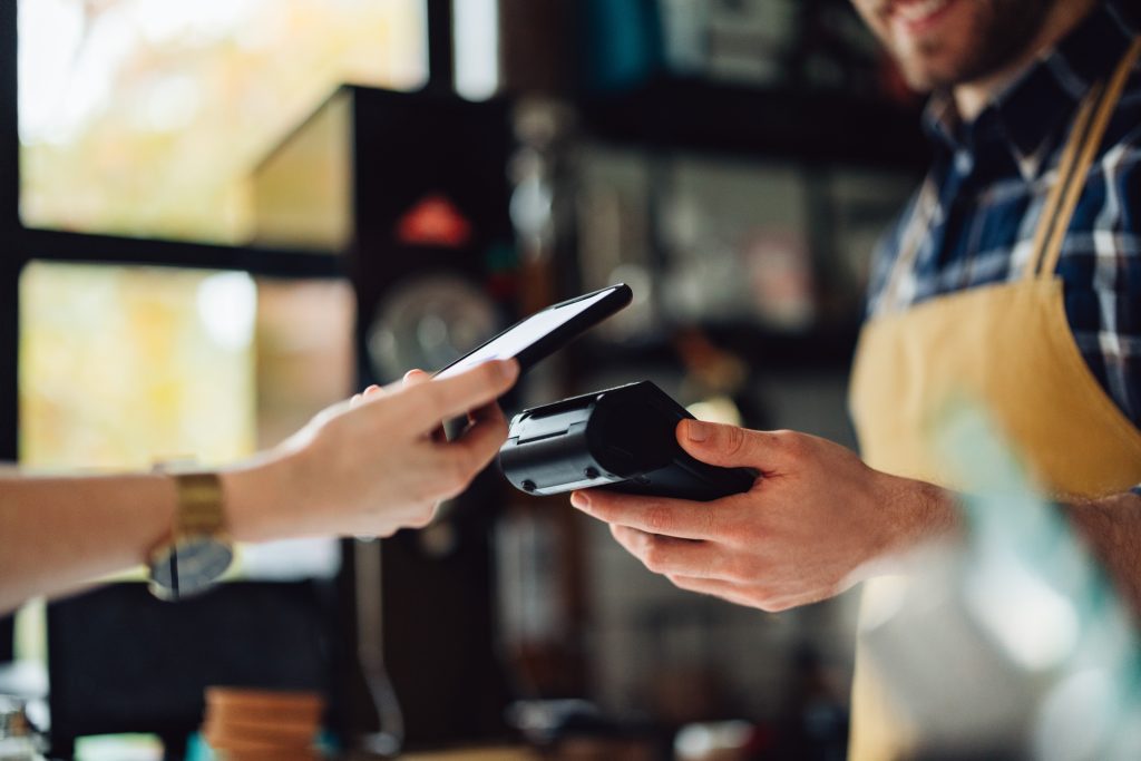 A customer making a contactless payment using a smartphone at a cafe, with the barista holding the payment terminal.