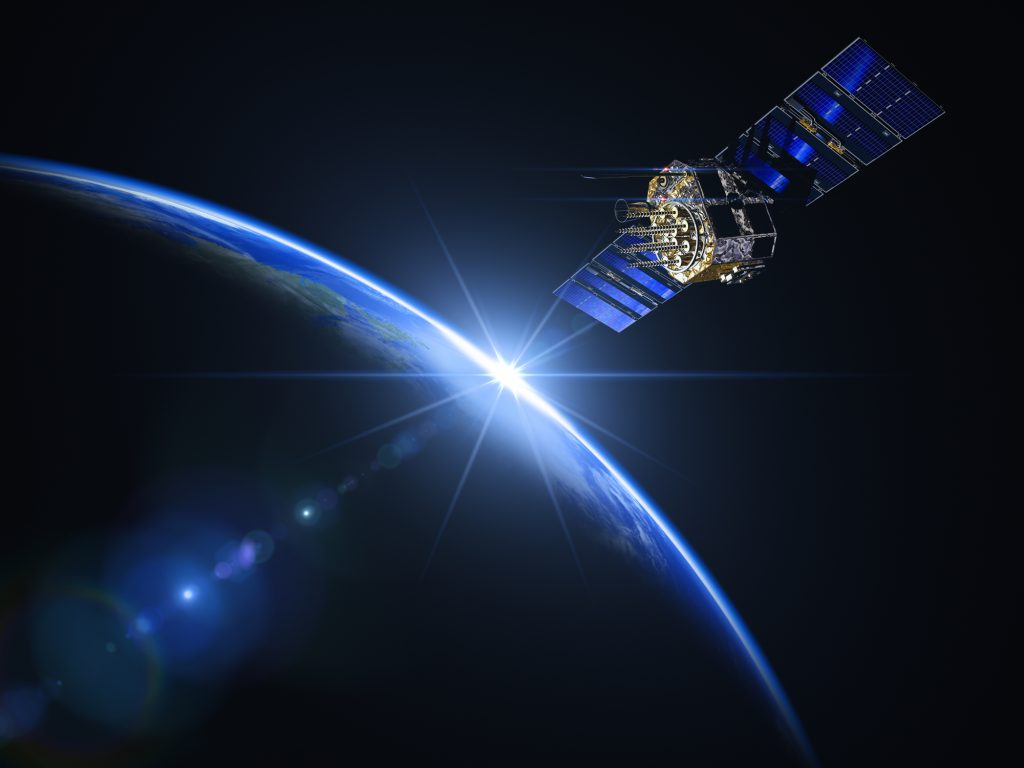 A satellite with solar panels orbits above Earth with the sun peeking over the horizon in the background.